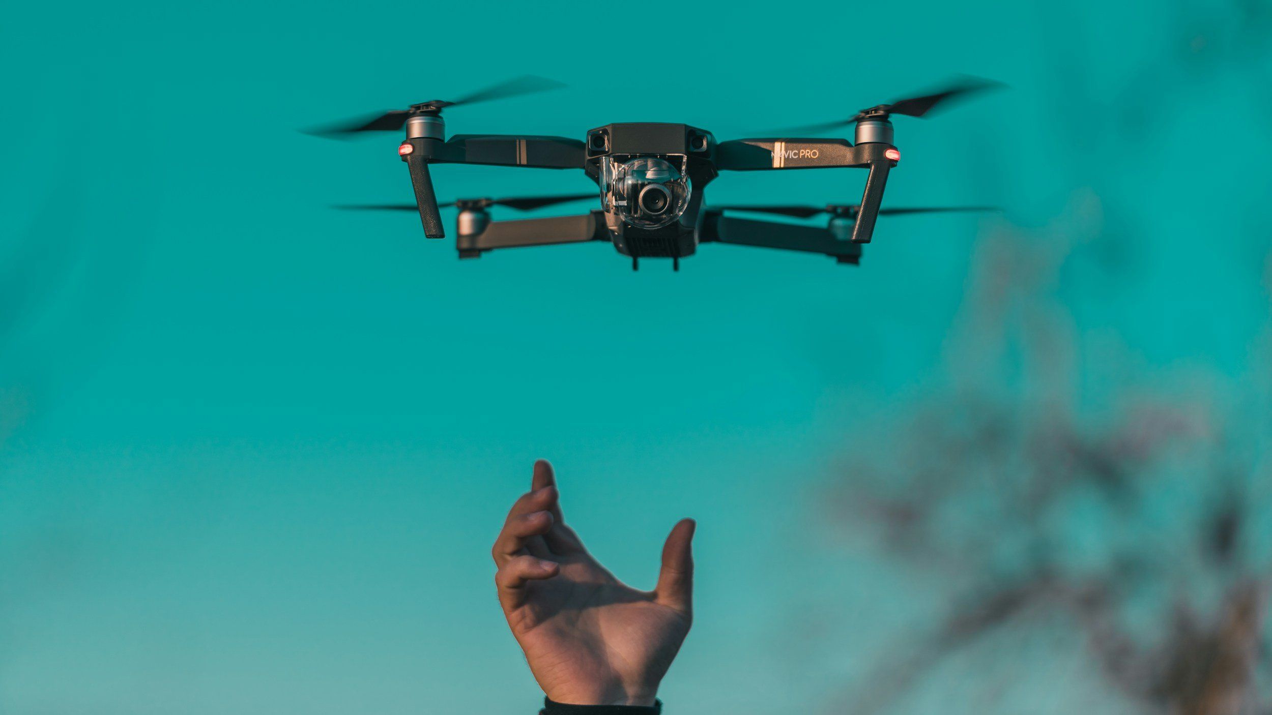 Camera drone hovering above an outstretched hand against a teal sky